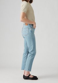 Lichtblauwe denim jeans met een rechte snit, voorzien van een ontwerpelement op de achterzak en een bruine leren patch. Gecombineerd met een beige top met korte mouwen en zwarte schoenen.