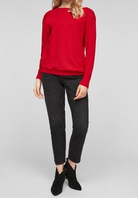 Red long-sleeved sweater with a textured pattern, paired with black slim-fit jeans and pointed black ankle boots. Neutral background.