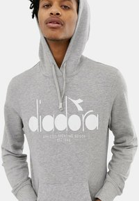 Felpa grigia con cappuccio, con un grande logo bianco che riporta la scritta "diadora" e "ATHLETES SPORTING GOODS EST. 1948" in carattere grassetto.
