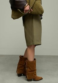 Jupe olive à mi-mollet associée à des bottines en suède marron avec des lacets, texture visible. Tenant un sac en olive texturé.