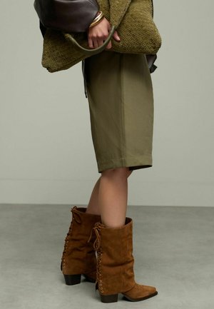 Jupe olive à mi-mollet associée à des bottines en suède marron avec des lacets, texture visible. Tenant un sac en olive texturé.