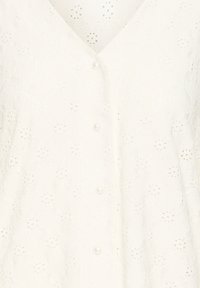 Blouse blanche avec un col en V et une fermeture à boutons, ornée de motifs floraux brodés et de petits boutons en perle. Tissu texturé.