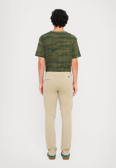 Grönmönstrad t-shirt med rund hals, kombinerad med beige slim-fit byxor. Skorna har en turkos accent och bruna sulor.