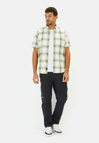 Korte mouwen geruit shirt in groen en wit, gecombineerd met marineblauwe cargobroeken met meerdere zakken. Draagt witte sneakers.