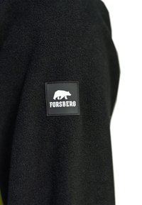 Schwarze Fleecejacke mit einem quadratischen Patch mit dem Wort "FORSBERG" und einem Bärenmotiv in Weiß. Weicher Stoff und einfaches Design.