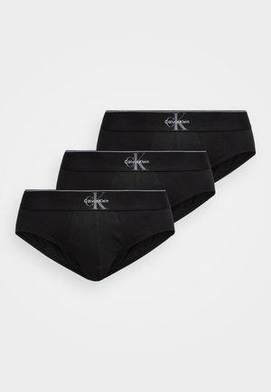 Drei schwarze Herren-Slips von Calvin Klein mit weißem Logo auf den elastischen Bundstreifen, auf einem grauen Hintergrund gestaffelt übereinander angeordnet.