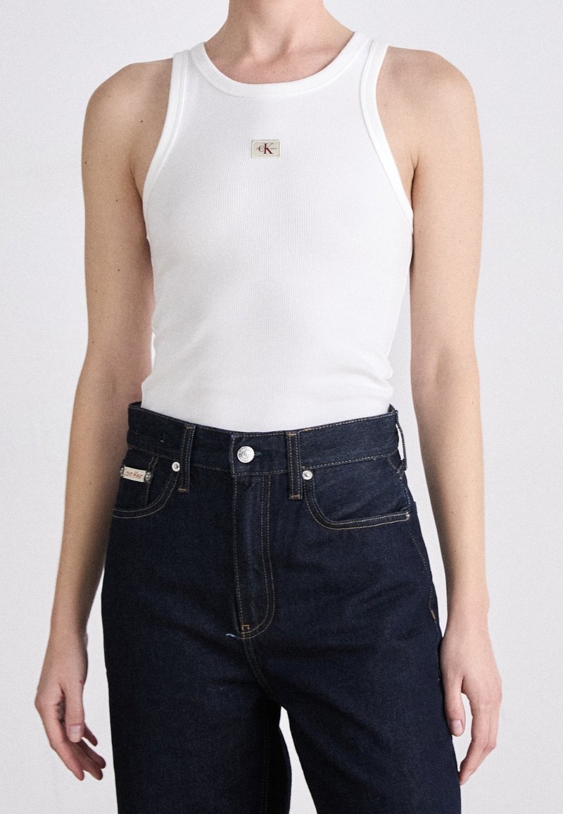 Weißes geripptes Tanktop mit einem kleinen CK-Logo-Patch, kombiniert mit dunkelblauen, hoch taillierten Jeans mit Metallknöpfen und Nähtelementen.