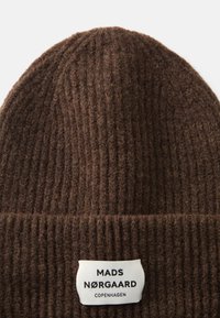 Beanie lavorato a maglia marrone con texture a coste, dotato di un'etichetta con la scritta "MADS NØRGAARD COPENHAGEN." Il materiale sembra morbido e caldo.