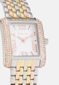 Zegarek Michael Kors z prostokątną bransoletą w kolorze srebrnym i złotym, akcentami w kolorze różowego złota, białą tarczą oraz cyframi rzymskimi jako znacznikami godzin.