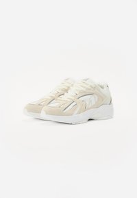 Beige en witte sneakers met een mesh bovenwerk, suède accenten, ronde neus en dikke zool. Kenmerken zijn een vetersluiting en contrasterende stiksels.