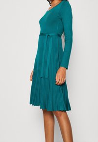 Robe turquoise, côtelée, avec des manches longues, un décolleté en V et une taille ceinturée. La jupe présente des plis verticaux, créant une silhouette fluide.