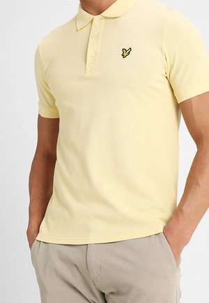 Mężczyzna ubrany w jasnżółtą koszulkę polo z krótkim rękawem i małym czarno-żółtym logo ptaka na piersi, w połączeniu z beżowymi spodniami.