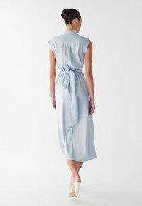 Hele sinise ilma käisteta kleit, millel on sidumisvöö ja keskmise sääre pikkus, valmistatud siledast kangast; omadused on krae ja peene tagumise lõhiku detail.