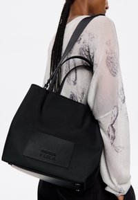 Borsa tote nera con tessuto morbido, doppio manico superiore, patch logo discreto e design strutturato, indossata sopra una blusa leggera a fantasia.