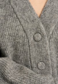 Pilkas kardiganas, pagamintas iš riboto audinio, turintis gilų V formos iškirptę, tekstūrinį raštą ir apvalių mygtukų akcentus. Minkšta, jauki tekstūra.