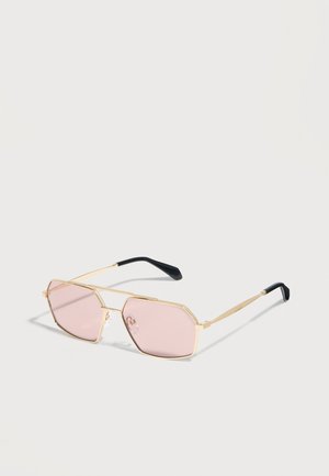 Lunettes de soleil à monture dorée avec des verres géométriques roses et des embouts d'oreille noirs reposant sur une surface blanche.