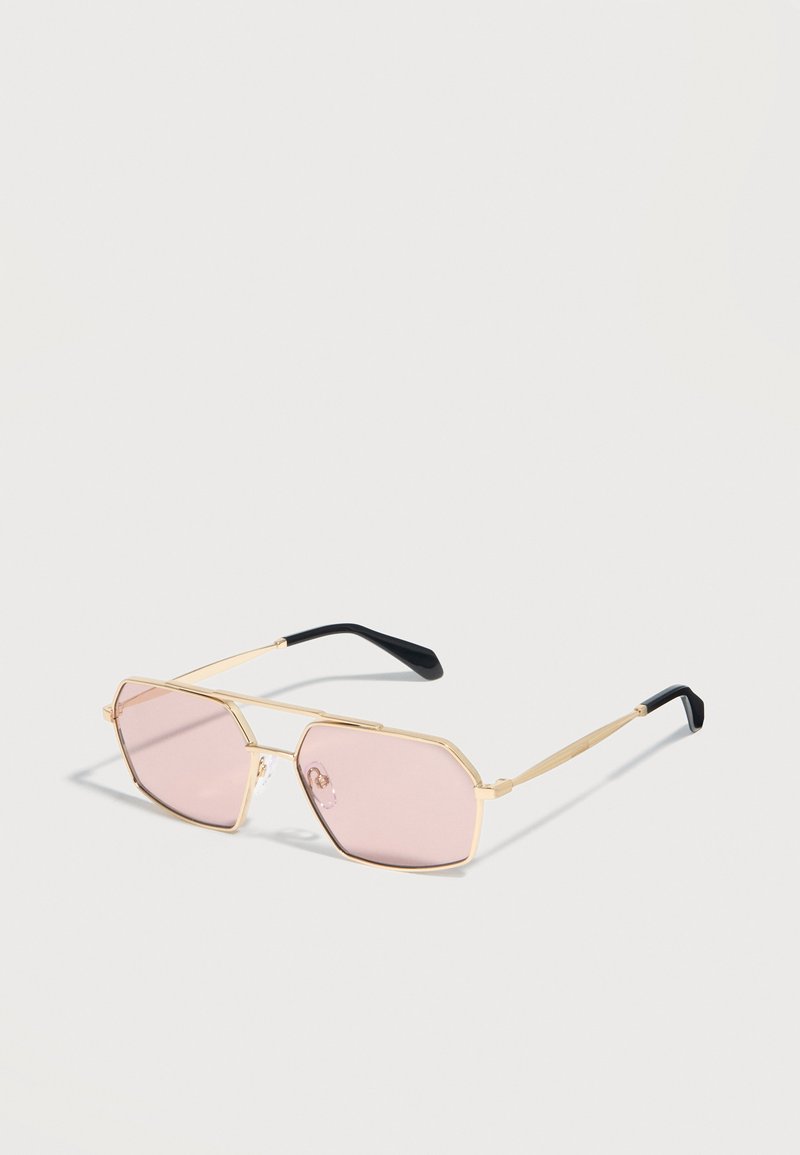 Γυαλιά ηλίου με χρυσή κορνίζα, γεωμετρικούς ροζ φακούς και μαύρα άκρα στα σκέλη, τοποθετημένα πάνω σε μια λευκή επιφάνεια.