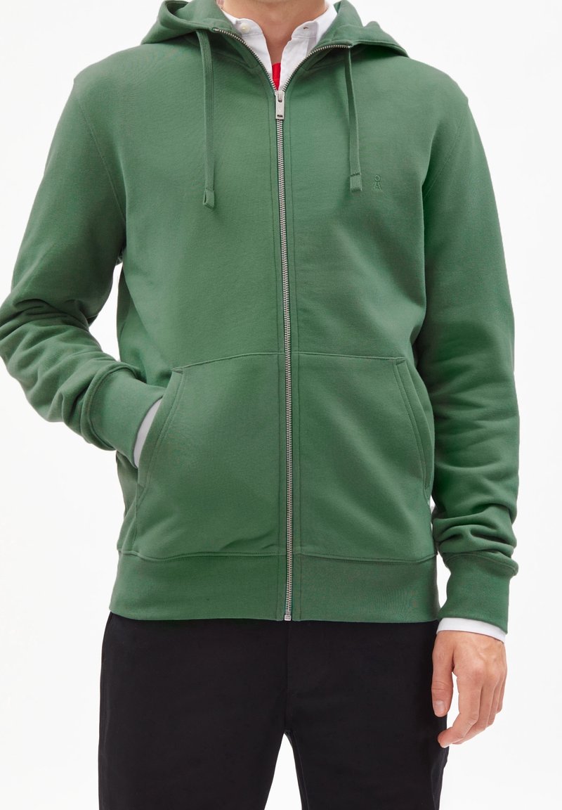 Homme portant un sweat à capuche vert zippé avec des poches avant, une chemise blanche en dessous, et un pantalon noir, debout devant un fond uni.
