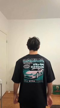 Melna lielizmēra t-krekls ar vēsturiska automobiļa grafiku rozā un tirkīza krāsā uz muguras, papildināts ar kontrastējošiem tekstiem un ģeometriskām rakstiem.