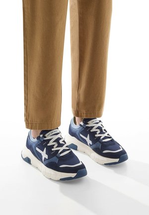 Marineblaue Sneakers aus einer Mischung aus Wildleder und Mesh-Material, mit weißen Akzenten und dicker Sohle. Kombiniert mit braunen Cropped-Hosen.