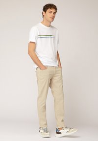 T-shirt bianca con strisce orizzontali verdi e blu, pantaloni beige e sneaker multicolori; fit casual, tessuto morbido e tonalità neutre.