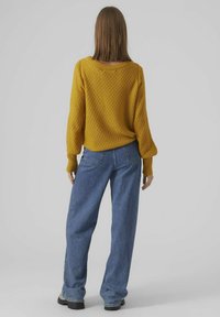 Gul stickad tröja med ett strukturerat mönster, vida ärmar och en avslappnad passform, kombinerad med högmidjade blå jeans.