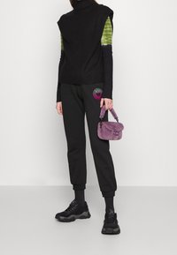 Czarny bezrękawnik, pod nim zielona i czarna koszula z długim rękawem. Czarnie dresy z kolorową grafiką. Fioletowa torebka z sztucznego futra, czarne sneakersy.