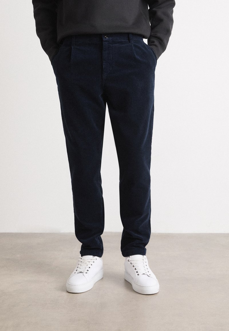Personne portant un pantalon en velours côtelé bleu marine foncé et des baskets blanches, debout sur un sol gris clair les mains dans les poches.