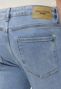 Close-up van een lichtblauwe denim jeans achterzak en een beige merklabel met tekst Marc O'Polo Denim bij een persoon die een wit geribd top draagt.