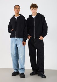 Sudaderas con cremallera negras llevadas sobre camisetas blancas, combinadas con jeans de pierna ancha en azul claro y oscuro, sobre un fondo liso.