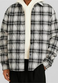 Vadderad flanellskjorta i svart och vit rutig mönster. Har tryckknappar, krage och en avslappnad passform, lagd över en lätt huvtröja.