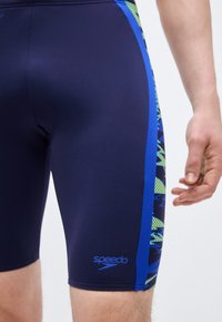 Άνδρας φοράει ναυτικό μπλε μαγιό Speedo jammers με πλευρικά πάνελ σε μπλε και πράσινο μοτίβο, στέκεται μπροστά σε απλό φόντο.