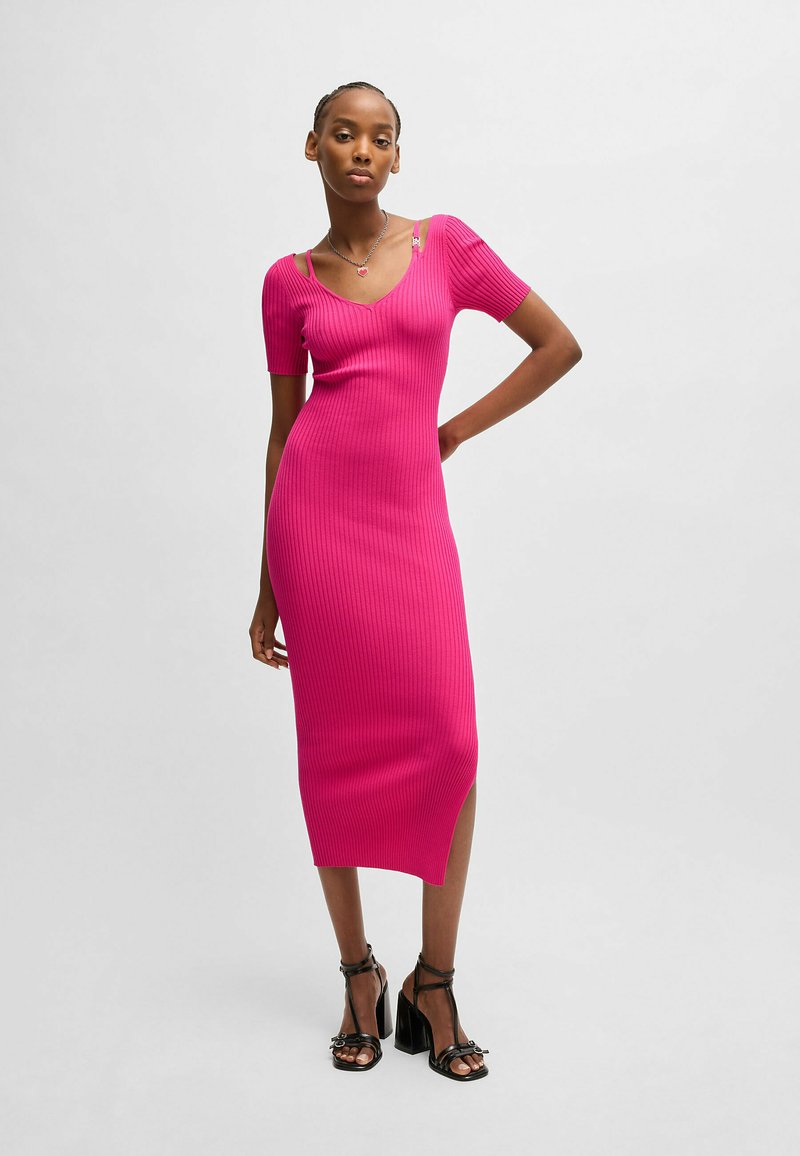 Dopasowana, prążkowana sukienka w intensywnym różu, z krótkimi rękawami, dekoltem w kształcie litery V oraz rozcięciem z boku, w połączeniu z czarnymi sandałkami na obcasie.