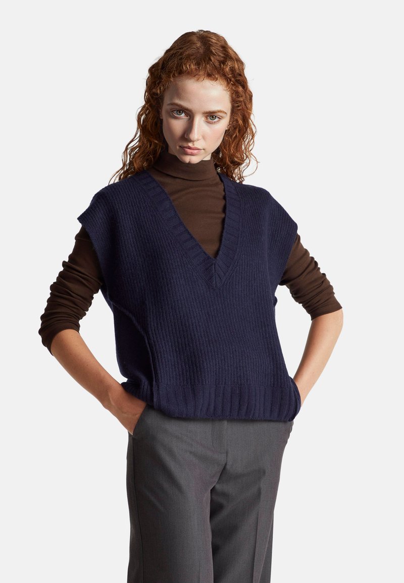 Gilet a costine blu navy con scollatura a V e maniche corte indossato sopra un dolcevita marrone, abbinato a pantaloni grigi.