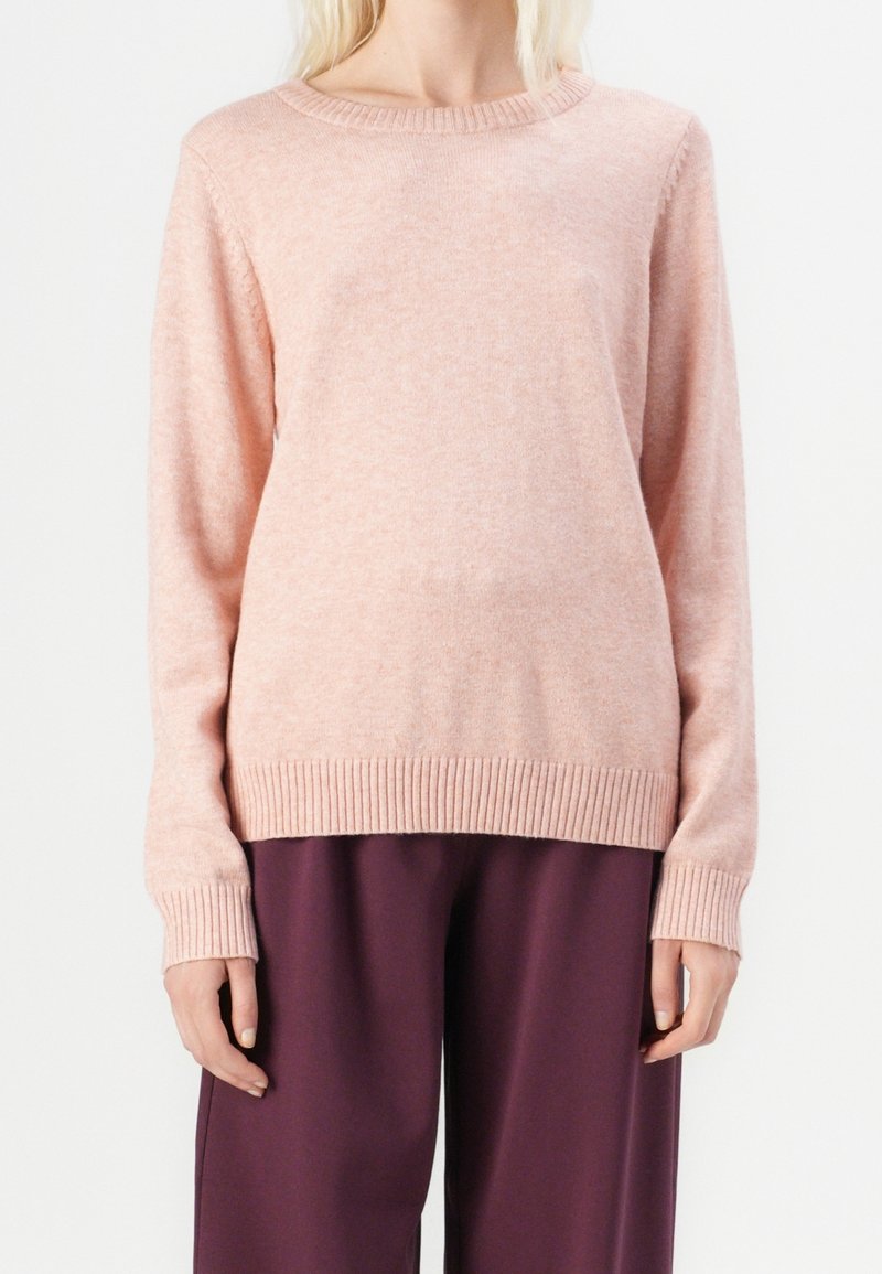 Pull rose clair en tricot avec col rond, poignets et ourlet côtelés. Tissu doux avec une finition légèrement texturée. Associé à un pantalon violet foncé.