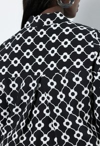 Czarna koszula z geometrycznym, białym wzorem kwiatowym połączonym liniami. Posiada klasyczny kołnierz i zakładkę z tyłu.