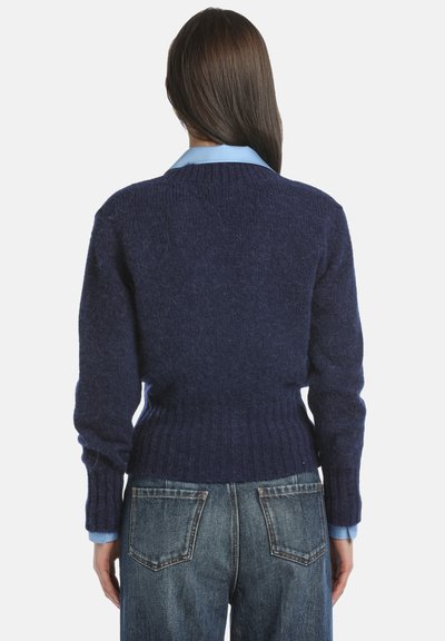 Mornarska bluza iz mehkega pletenega materiala, z ribasto obrobo in manšetami, oblečena čez svetlo modro srajco s kratkimi rokavi, v kombinaciji z jeans hlačami.