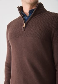Pullover marrone lavorato a maglia con colletto alto, dotato di zip frontale a un quarto e dettagli in pannello testurizzati. Tessuto liscio con accenti a coste.