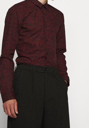 Mann, der ein eng anliegendes, dunkelrotes und schwarzes Hemd mit Muster trägt, in die hoch taillierte schwarze Hose gesteckt, mit den Händen in den Taschen.