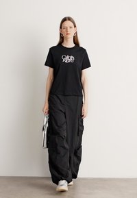 T-shirt noir en coton avec logo blanc, associé à un pantalon cargo noir ample doté de poches et d'une finition texturée. Sneakers argentées et sac métallique.
