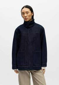 Giacca di jeans scuro con collo alto, tasche frontali, taglio dritto e cuciture visibili. Indossata sopra pantaloni di colore chiaro.