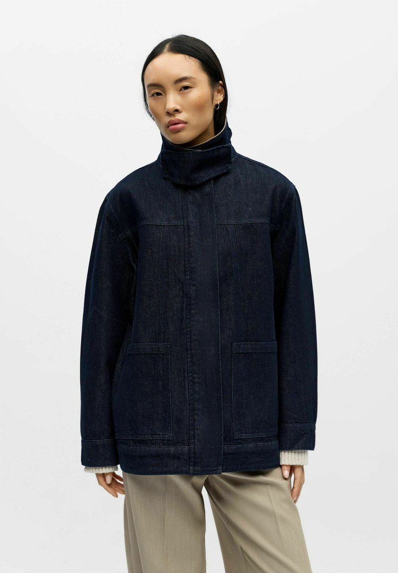 Giacca di jeans scuro con collo alto, tasche frontali, taglio dritto e cuciture visibili. Indossata sopra pantaloni di colore chiaro.