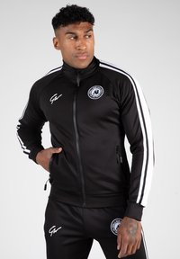 Zwarte zip-up trainingsjack met witte strepen, zijzakken, ronde kraag en logo op de borst. Stof lijkt glad en aansluitend.