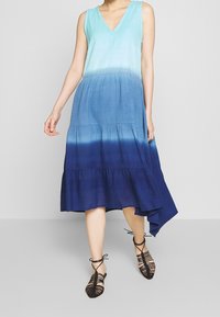Une robe sans manches à col en V, avec un dégradé allant du bleu sarcelle clair au bleu marine profond, ornée de volants superposés et d'une coupe décontractée, fabriquée à partir d'un tissu léger.