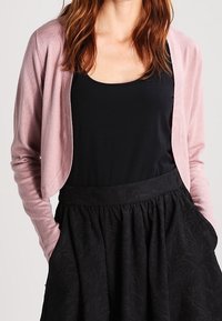 Femme portant un cardigan rose clair, un haut sans manches noir et une jupe noire taille haute, les mains dans les poches, devant un fond uni.