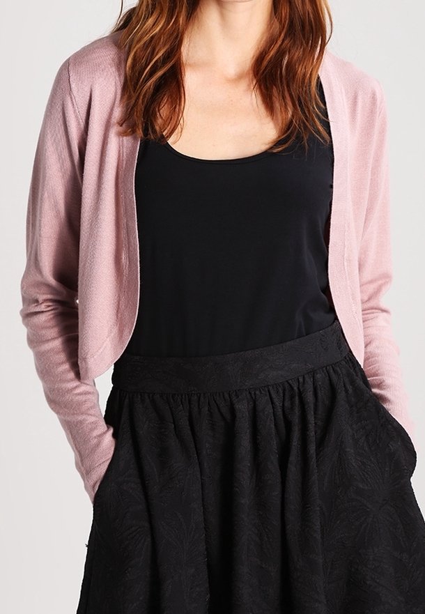 Femme portant un cardigan rose clair, un haut sans manches noir et une jupe noire taille haute, les mains dans les poches, devant un fond uni.