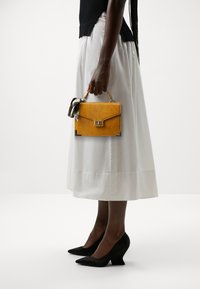 Keltainen krokotiilikuvioitu käsilaukku, jossa yläkahva ja koristeellinen huivi, yhdistetty valkoiseen hameeseen ja mustiin kiilakorkoihin.