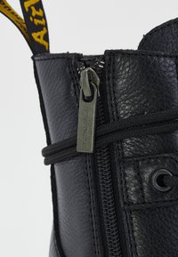 Black leather ankle boot featuring a side zipper, textured surface, and black lacing. Includes a yellow branded pull tab and reinforced stitching.