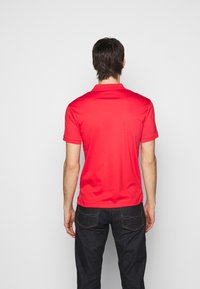 Rotes Polohemd mit kurzärmligen, einem Kragen und glattem Stoff. Tragen mit dunklen Jeans, von hinten betrachtet. Minimale Details.