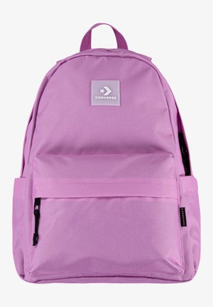 Mochila de tela rosa con un bolsillo frontal con cremallera, que presenta un parche con el logo de Converse y un asa superior. Textura suave, forma clásica.